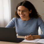 A young woman with curly brown hair and glasses smiles while working at her laptop in a sunlit home office. She wears a soft blue sweater and writes in a notebook beside a coffee mug and pencil cup, with a cozy, plant-filled background.